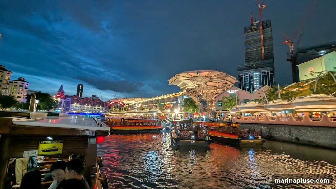 A wide shot of a riverfront at dusk, with several tour boats docked and moving on the water. Buildings line the riverbank, illuminated by lights, with a modern skyscraper under construction in the background. The sky is a deep blue with dramatic clouds.