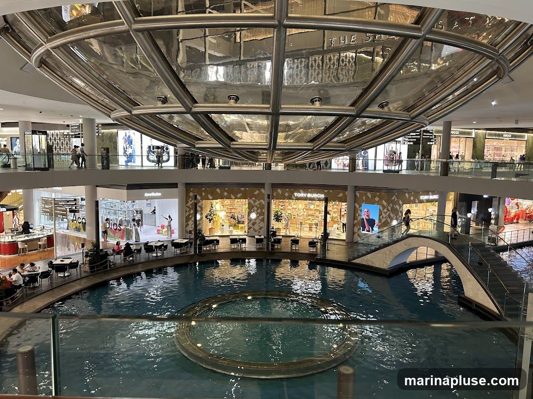 An interior view of a multi-level shopping mall featuring a large, circular water feature in the center. Shops are visible on multiple levels, including Tory Burch and Gucci. People are seen walking on elevated walkways and bridges, and seated at tables near the water. A large, reflective, dome-like structure hangs from the ceiling.