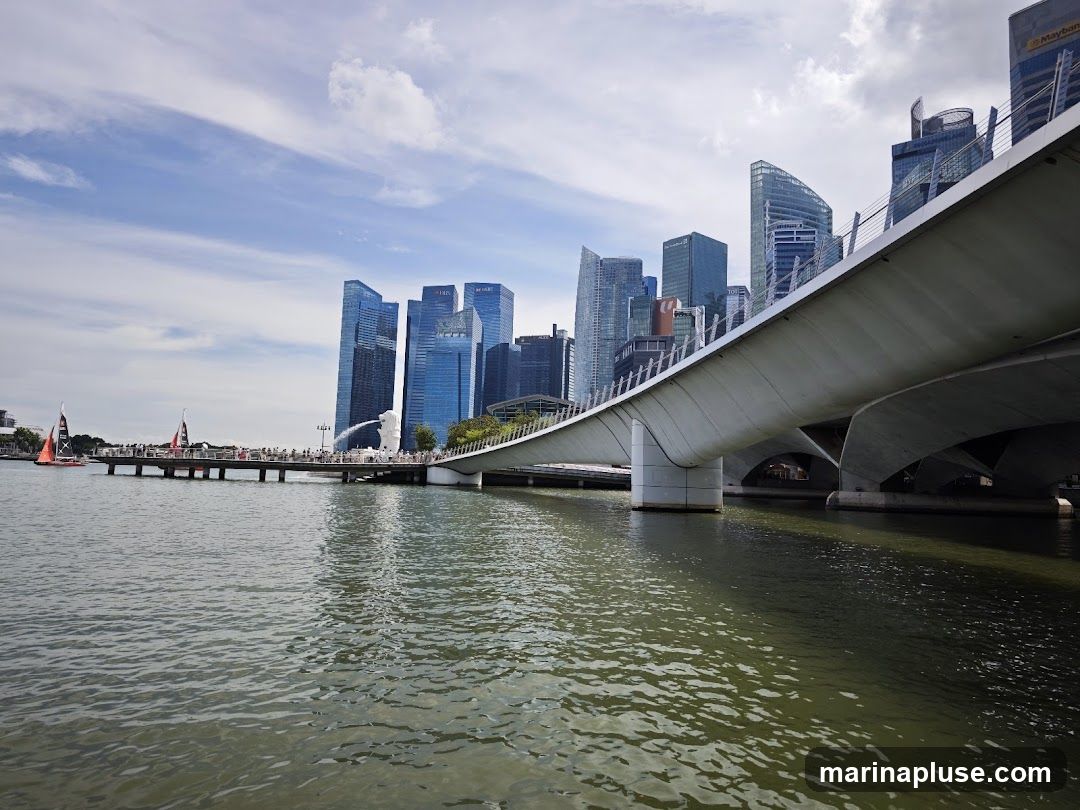 A wide shot of the Singapore skyline featuring modern skyscrapers and the iconic Marina Bay Bridge. The water in the foreground reflects the sky and buildings. Two sailboats are visible in the distance on the left side of the image, near a pier with people. The Merlion statue is also visible on the pier.