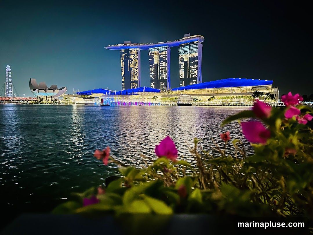 A nighttime view of the Marina Bay Sands complex in Singapore, with its iconic three towers illuminated and connected by a sky-high platform. The building's facade displays a grid of lit windows. In front of the towers, a long, low-rise structure with a bright blue roof is visible, also illuminated. The complex is situated across a body of water, which reflects the lights. In the foreground, out-of-focus pink flowers and green foliage add a touch of nature to the urban landscape. To the left, a Ferris wheel and a distinctive boat-shaped structure are also visible in the distance.