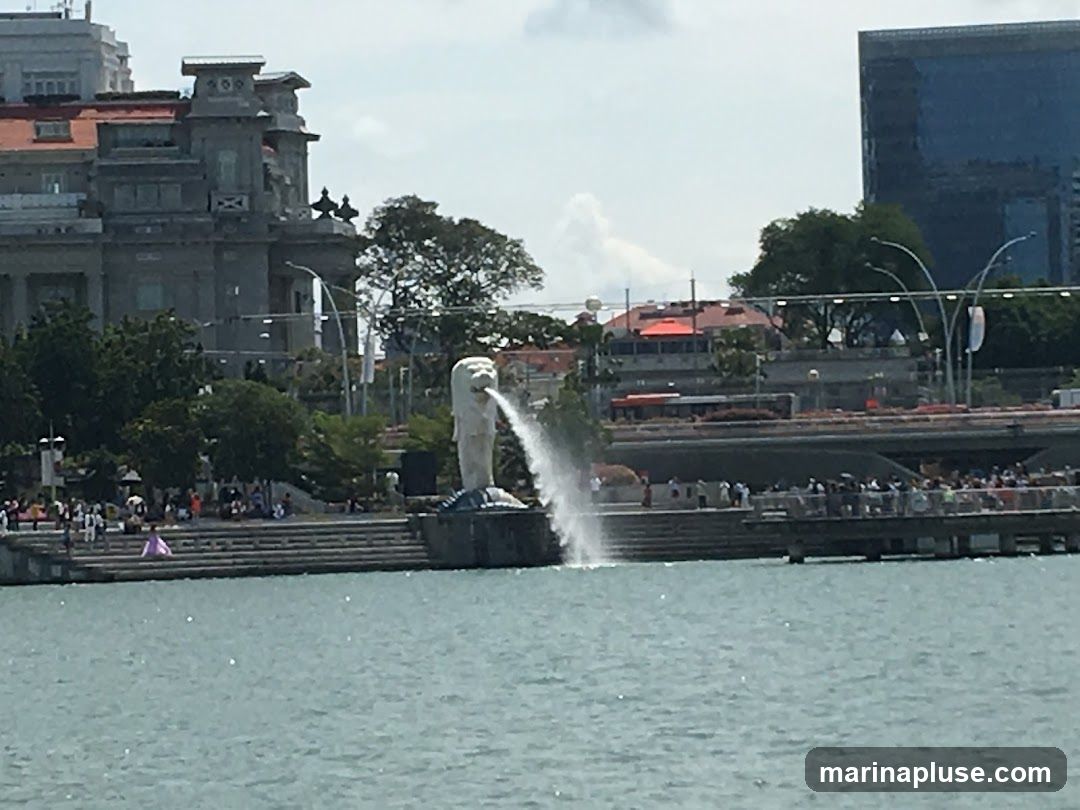 A wide shot of the Merlion statue in Singapore, spewing water into the sea. In the background, there are several buildings, including a modern skyscraper and a more traditional-looking structure. A bridge with traffic and a promenade with many people are also visible. The water is a calm blue, and the sky is partly cloudy.