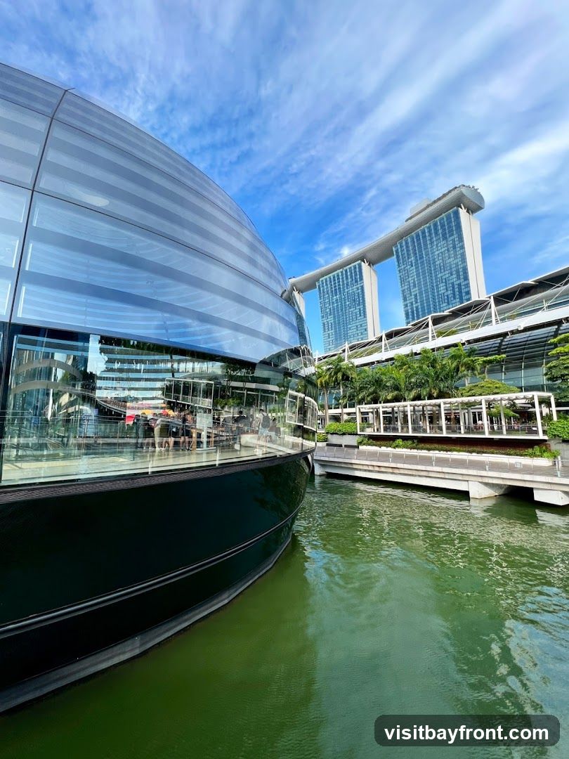 A modern building with a curved glass facade is reflected in green water. In the background, the iconic Marina Bay Sands hotel complex is visible under a blue sky with scattered clouds. Palm trees and other greenery line the waterfront.