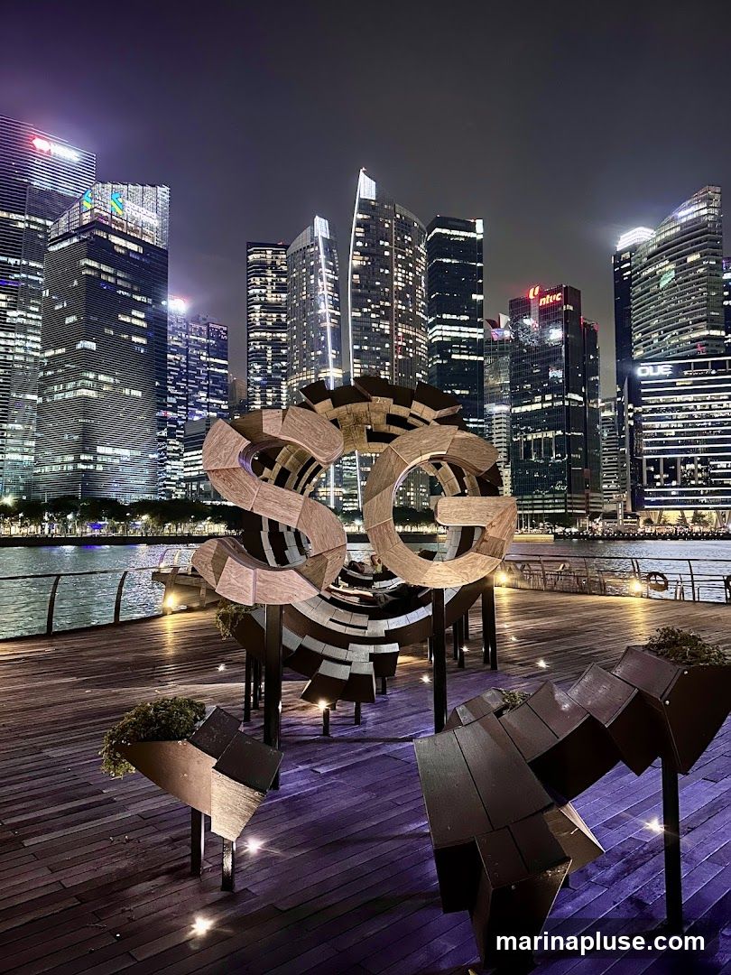 A nighttime view of the Singapore skyline with a large wooden sculpture in the foreground. The sculpture appears to be made of curved wooden planks and is illuminated by spotlights. The background features numerous illuminated skyscrapers and a body of water.