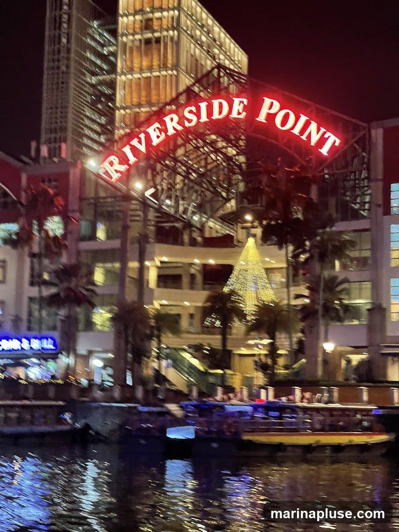 A neon sign reading "RIVERSIDE POINT" arches over a waterfront area at night. Buildings with illuminated windows and palm trees are visible in the background. Several boats are docked on the water, and the reflections of lights shimmer on the surface. A large, decorated Christmas tree is also visible.