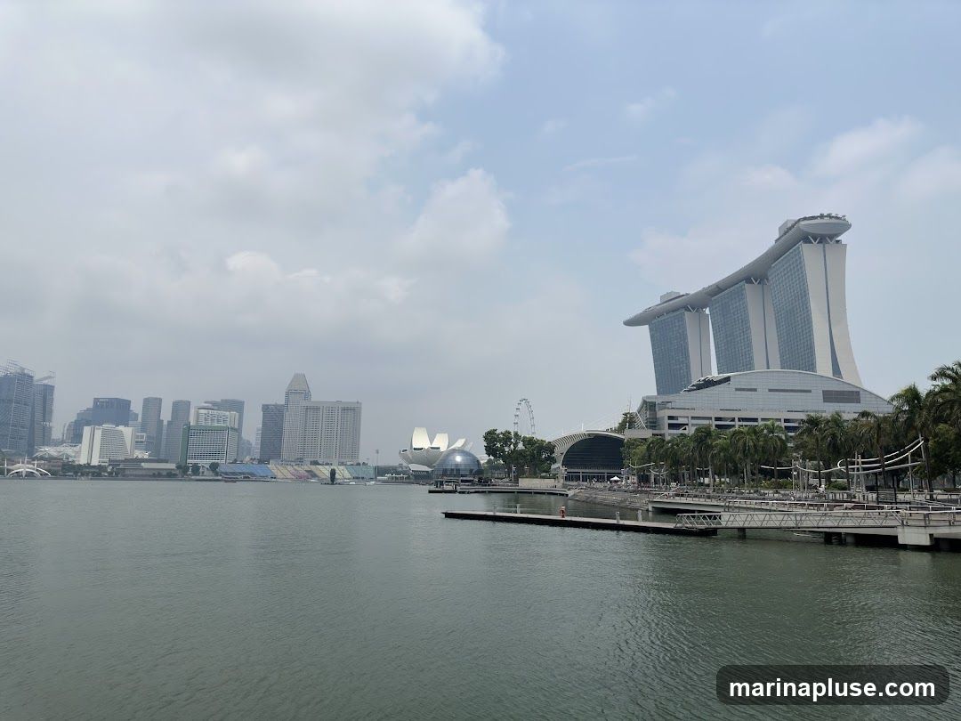 A wide shot of the Marina Bay area in Singapore, featuring the iconic Marina Bay Sands hotel with its distinctive boat-shaped top. In the background, the city skyline is visible, including various skyscrapers and the Singapore Flyer Ferris wheel. The foreground shows a body of water, likely Marina Bay itself, with a pier and walkway. The sky is partly cloudy.