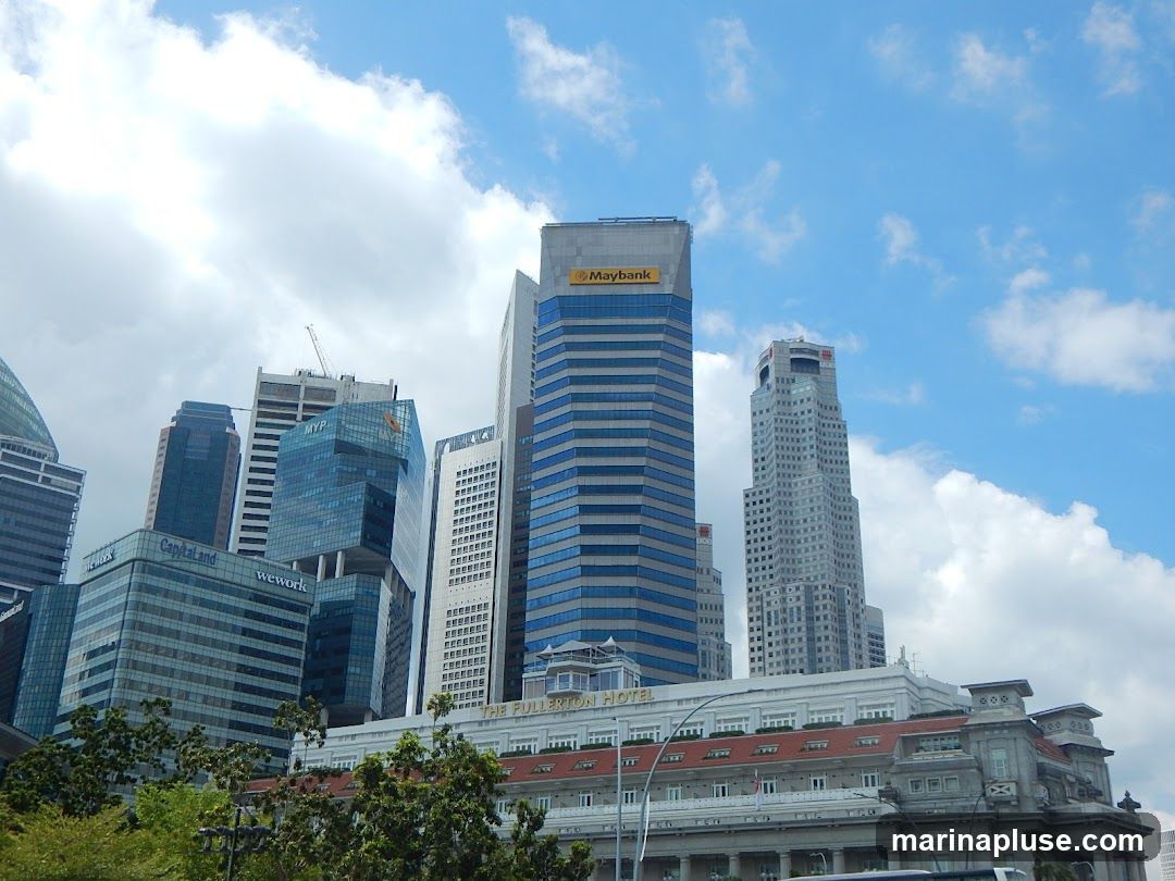 A view of a city skyline featuring several modern skyscrapers under a partly cloudy blue sky. Prominent buildings include those with signage for Maybank, CapitaLand, and WeWork. In the foreground, a lower, more traditional building with red-tiled roofs is visible, identified as The Fullerton Hotel. Lush green trees partially obscure the lower portion of the scene.