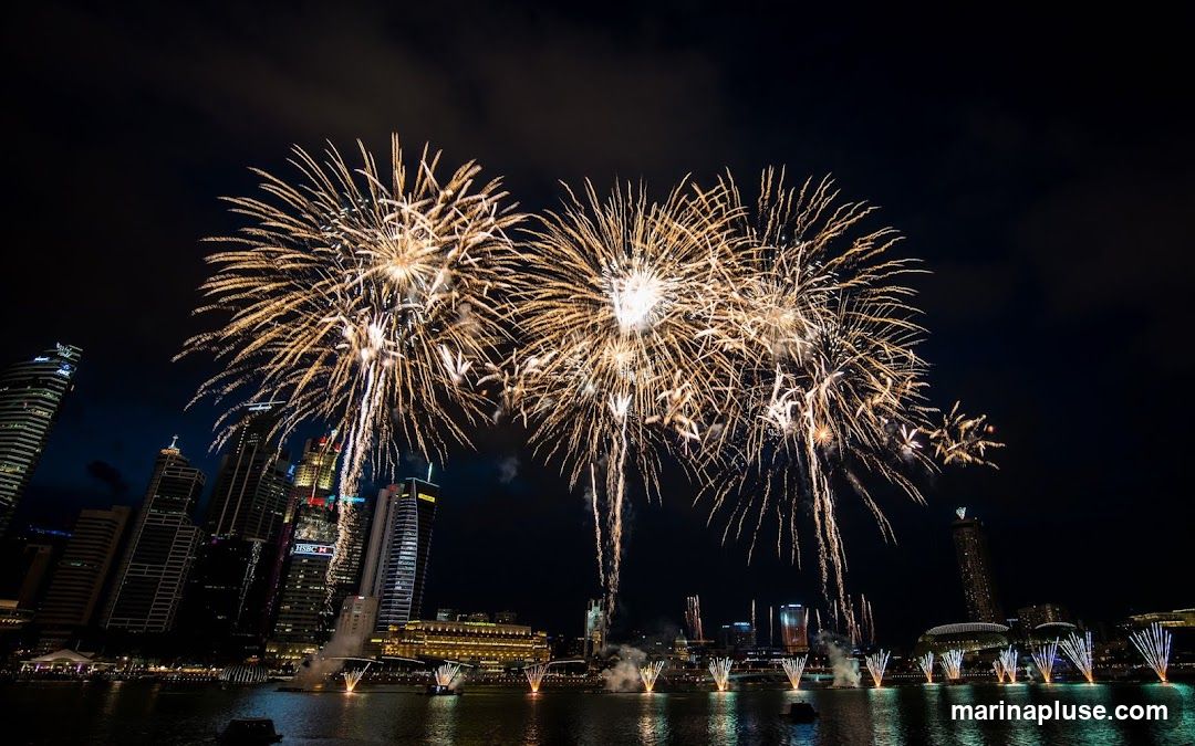A vibrant fireworks display illuminates the night sky above a modern city skyline, with numerous skyscrapers and buildings visible along the waterfront. The fireworks are predominantly golden and white, exploding in various patterns and sizes, casting a warm glow on the scene. Below, the city lights reflect on the calm water, and several boats or barges are positioned in the bay, some emitting plumes of smoke or light. The overall atmosphere is festive and celebratory.