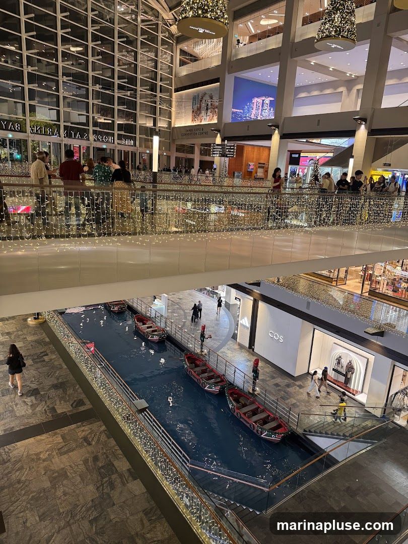 An interior view of a large, multi-level shopping mall. A canal with several red boats is visible on a lower level. People are walking on the upper levels and along the railings. The mall is decorated with festive lights. Stores like COS and Adidas are visible.