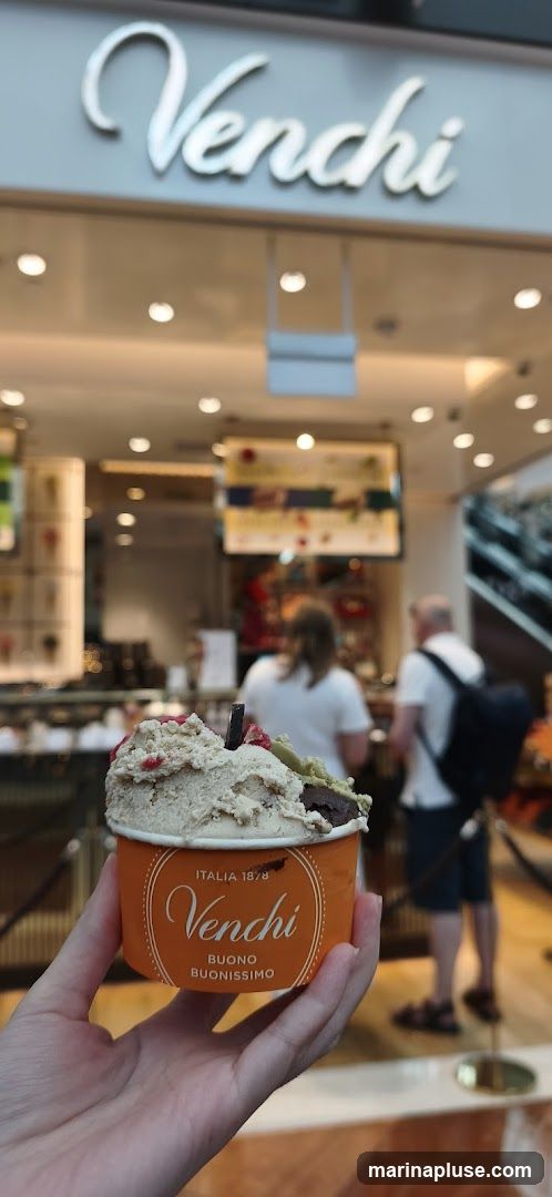 A hand holds an orange cup filled with gelato. The cup has the Venchi logo and "ITALIA 1878 BUONO BUONISSIMO" printed on it. In the background, the Venchi shop front is visible with its signage and interior.