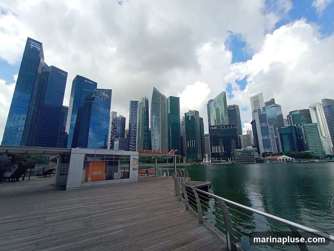 A wide shot of the Singapore skyline with its modern skyscrapers, viewed from a wooden boardwalk along the water. The sky is partly cloudy with patches of blue. In the foreground, a small modern structure with an orange panel is visible. The water reflects the buildings and the sky.