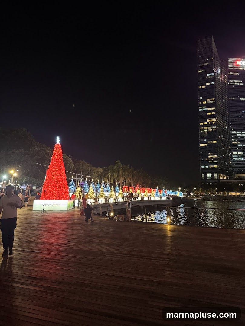 A nighttime scene featuring a large red Christmas tree illuminated with lights, followed by a row of smaller, colorful Christmas trees. People are walking along a wooden boardwalk next to a body of water. In the background, modern skyscrapers are visible, with some windows lit up. The overall atmosphere suggests a festive holiday event.
