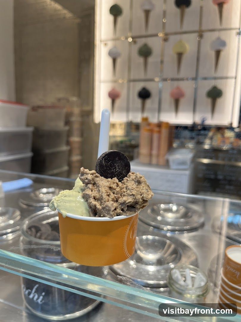 A close-up shot of a cup of gelato with two flavors, one light green and the other a chocolatey brown, topped with a dark cookie. The cup is orange and has a logo on the side. In the background, a display shows various ice cream cone designs.