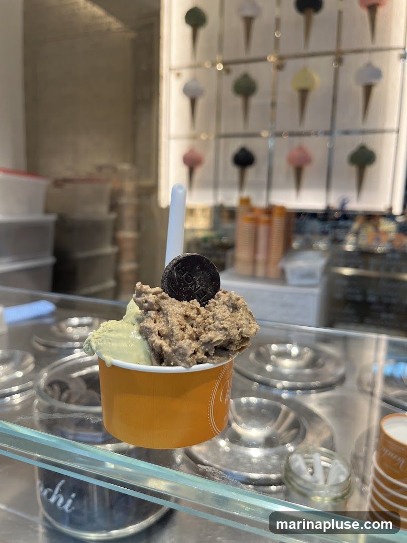 A close-up shot of a cup of gelato with two flavors, one light green and the other a chocolatey brown, topped with a dark cookie. The cup is orange and has a logo on the side. In the background, a display shows various ice cream cone designs.
