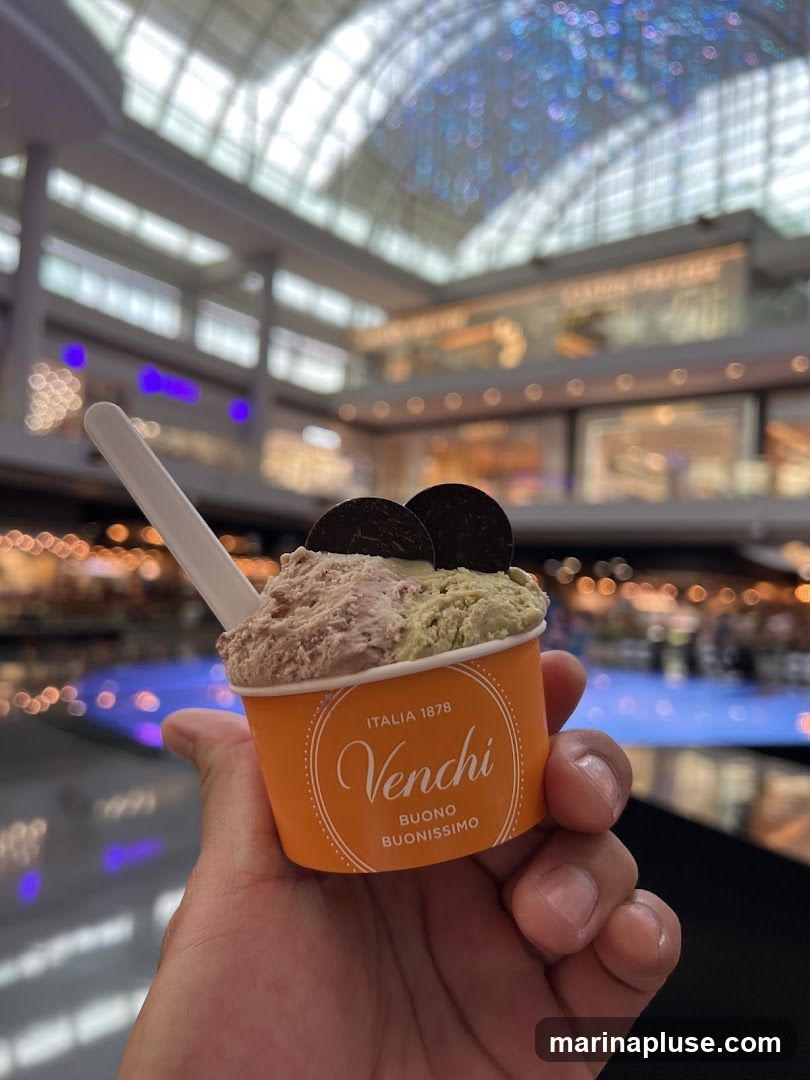 A hand holds an orange Venchi ice cream cup filled with two flavors of gelato, topped with two chocolate cookies. The background shows a blurred interior of a modern shopping mall with a glass ceiling and various shops.