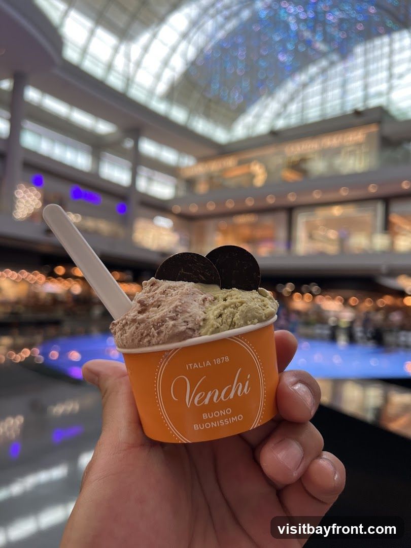 A hand holds an orange Venchi ice cream cup filled with two flavors of gelato, topped with two chocolate cookies. The background shows a blurred interior of a modern shopping mall with a glass ceiling and various shops.