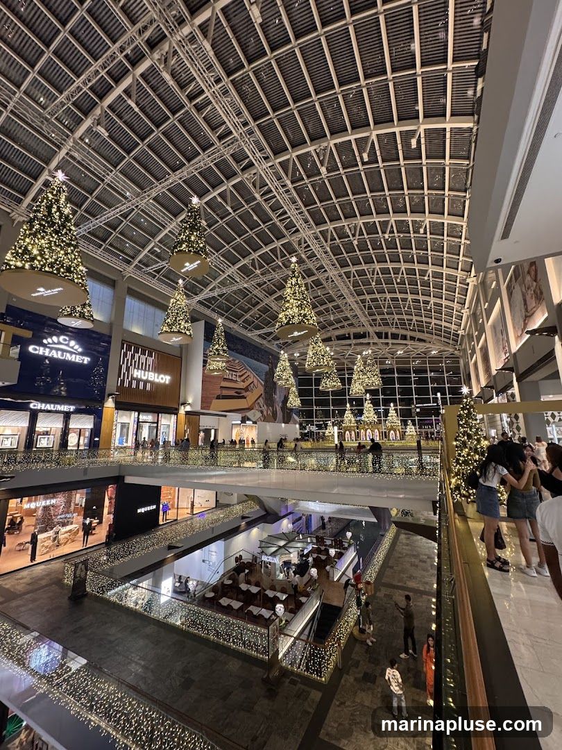 A wide shot of the interior of a shopping mall decorated for Christmas. Several Christmas trees adorned with lights are suspended from the ceiling. The mall features multiple levels with shops and escalators. People are seen browsing and walking around. The overall ambiance is festive and commercial.