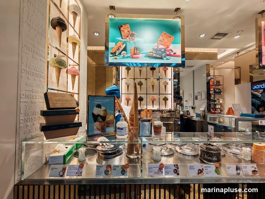 An interior view of a Venchi gelato shop, showcasing a display of gelato flavors, boxed chocolates, and decorative ice cream cones. The counter features various gelato tubs and a selection of packaged treats. A digital screen displays promotional images of Venchi products.