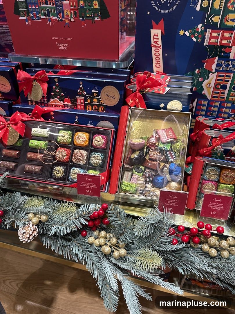 A display of Venchi chocolates in festive packaging, with Christmas trees and buildings depicted on the boxes. Several boxes are open, revealing an assortment of individually wrapped chocolates. A price tag of $85 is visible for one box, and another tag shows $40. The display is adorned with artificial pine garland, red berries, and gold decorative elements.