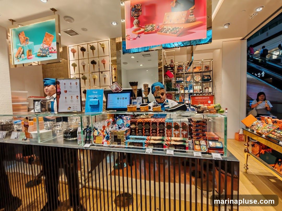A view inside a brightly lit shop selling chocolates and ice cream. Display cases are filled with various chocolate boxes and tubs of ice cream. Two staff members in blue hats are visible behind the counter. Digital screens show product information and prices. In the background, a display of ice cream cones is mounted on the wall, and escalators with people are visible to the right.