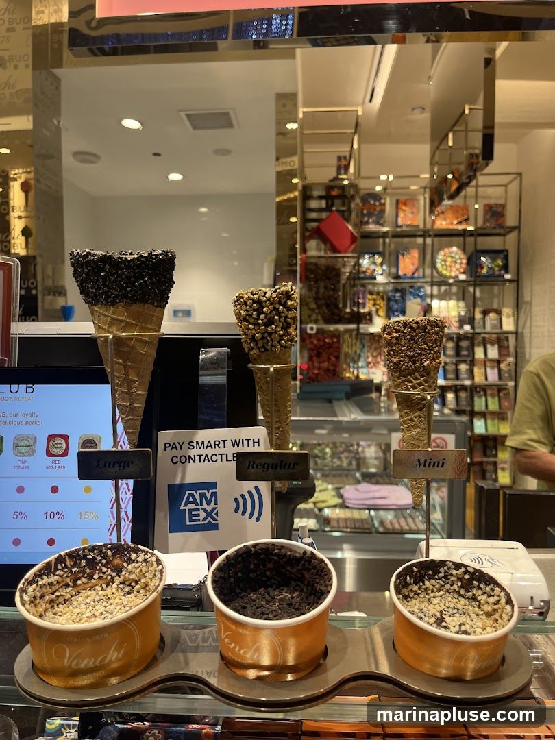 A display of ice cream cones and cups at a Venchi store. There are several cones with various toppings, including chocolate sprinkles and nuts. Below the cones are cups filled with ice cream and toppings. A sign indicates payment options and loyalty program tiers. In the background, shelves are stocked with chocolates and other confectionery items.