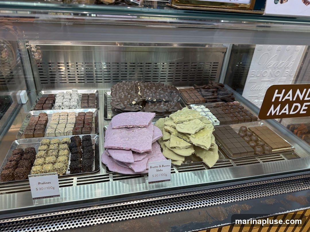 A display case filled with various types of chocolates, including pralines, chocolate bars, and chocolate slabs. Some chocolates are covered in nuts or other toppings. There are price labels for some of the items. A sign in the background reads "HAND MADE".