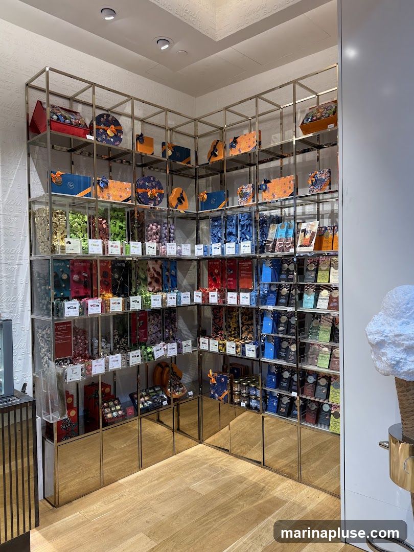A well-lit interior view of a shop showcasing a wide variety of chocolates and confections arranged on metal shelving units. The shelves are stocked with colorful boxes, individually wrapped chocolates, and jars of sweets. The walls are decorated with a repeating pattern that appears to be a brand logo or name. The flooring is light-colored wood. A large, abstract sculpture resembling an ice cream cone is visible on the right side of the image.