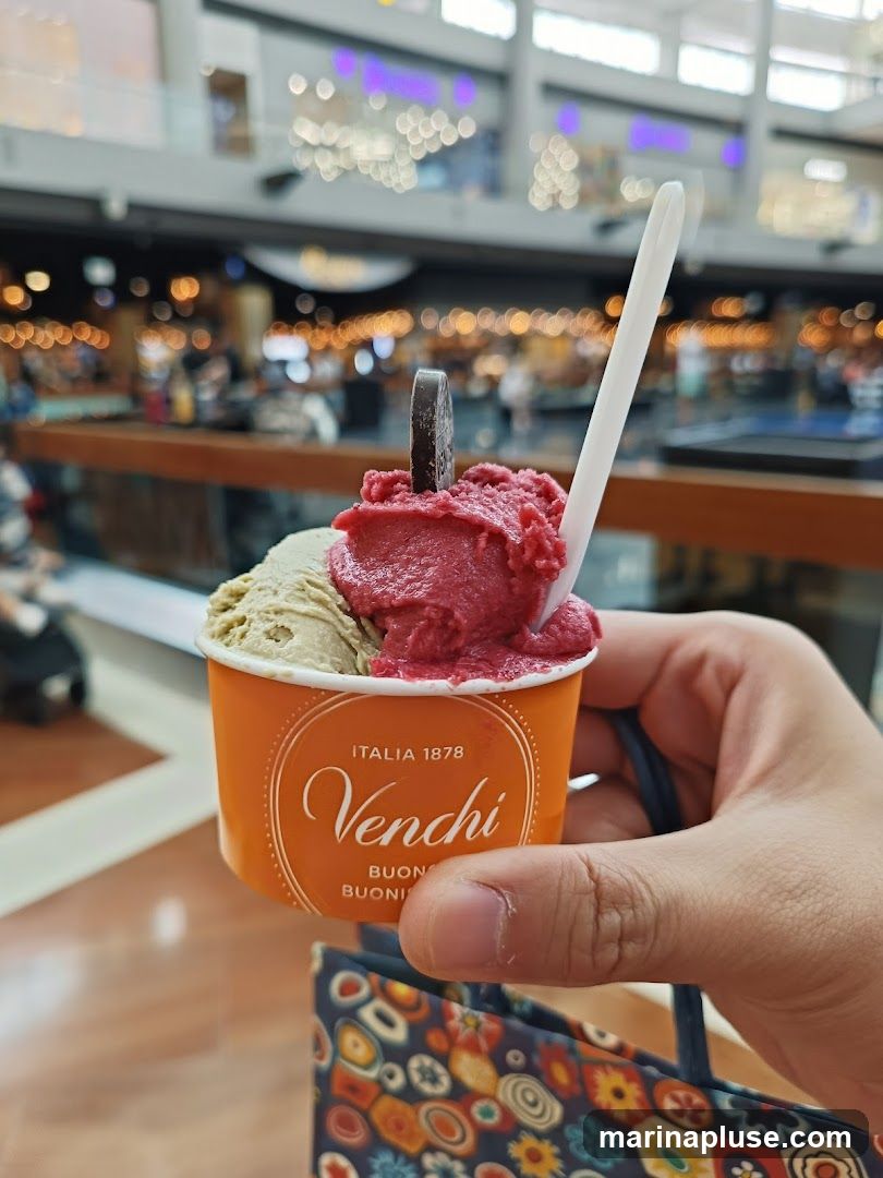 A hand holds an orange Venchi gelato cup filled with two scoops of gelato, one pistachio green and the other a deep red berry flavor, topped with a dark chocolate piece and a white spoon. The cup has the Venchi logo and "ITALIA 1878" printed on it. In the background, a blurred view of a shopping mall interior with shops and lights is visible. A patterned shopping bag is also partially visible.