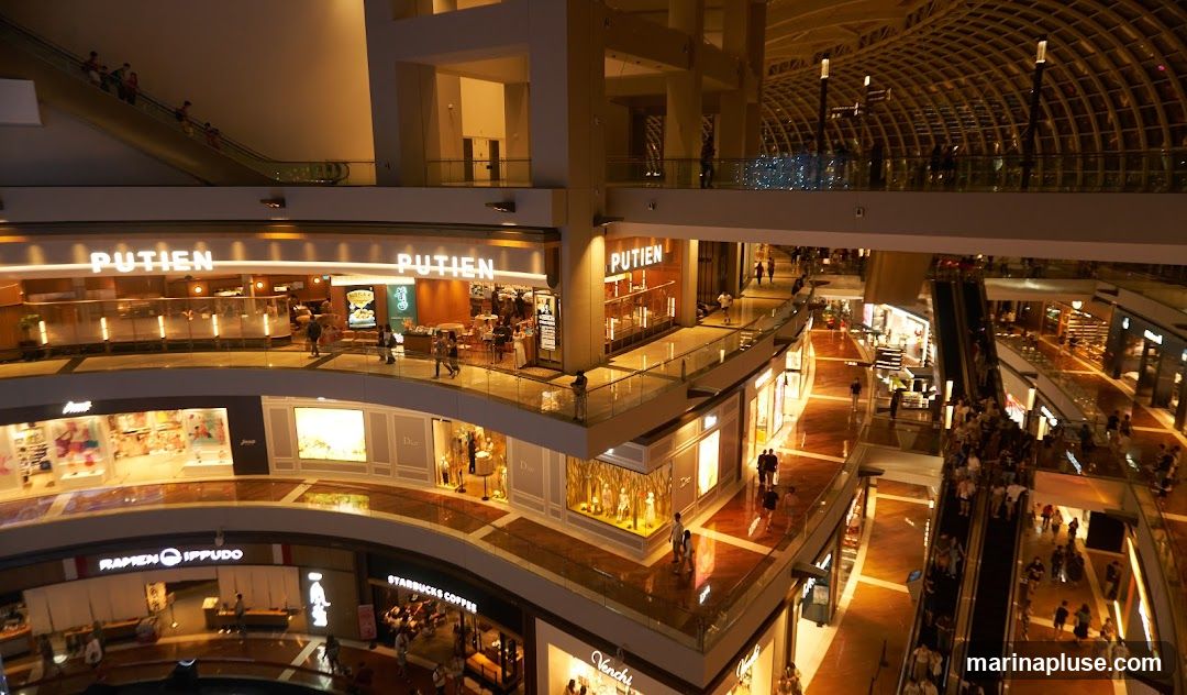 An interior view of a bustling shopping mall with multiple levels. Escalators and walkways connect different floors, with numerous shops visible. The architecture features a modern design with a curved, lattice-like ceiling. People are seen shopping, walking, and using the escalators. Prominent shop signs like "PUTIEN", "Dior", "Ramen Ippudo", and "Starbucks Coffee" are visible.