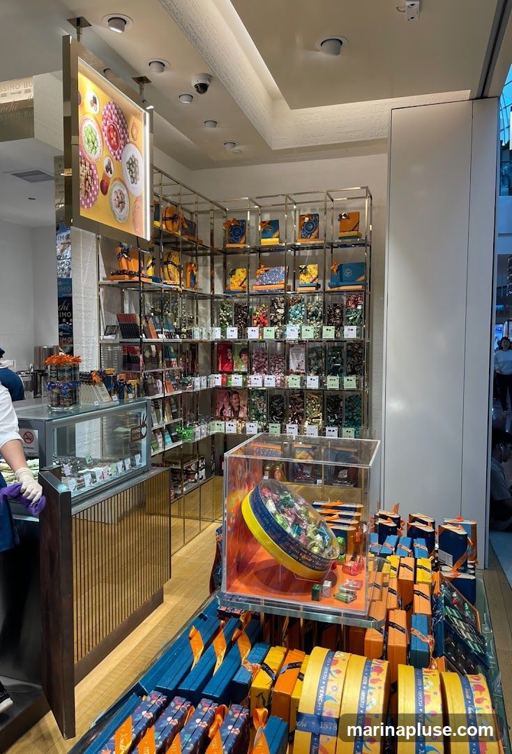A well-lit interior view of a shop showcasing a wide array of chocolates and confections. Shelves are filled with colorful boxes and individual chocolates in clear containers. A display case in the foreground holds a large, round tin overflowing with wrapped chocolates, surrounded by neatly stacked gift boxes. Another display case to the left shows smaller desserts or chocolates. The shop's branding, including the name "VENCHI," is visible on some of the packaging and ribbons.