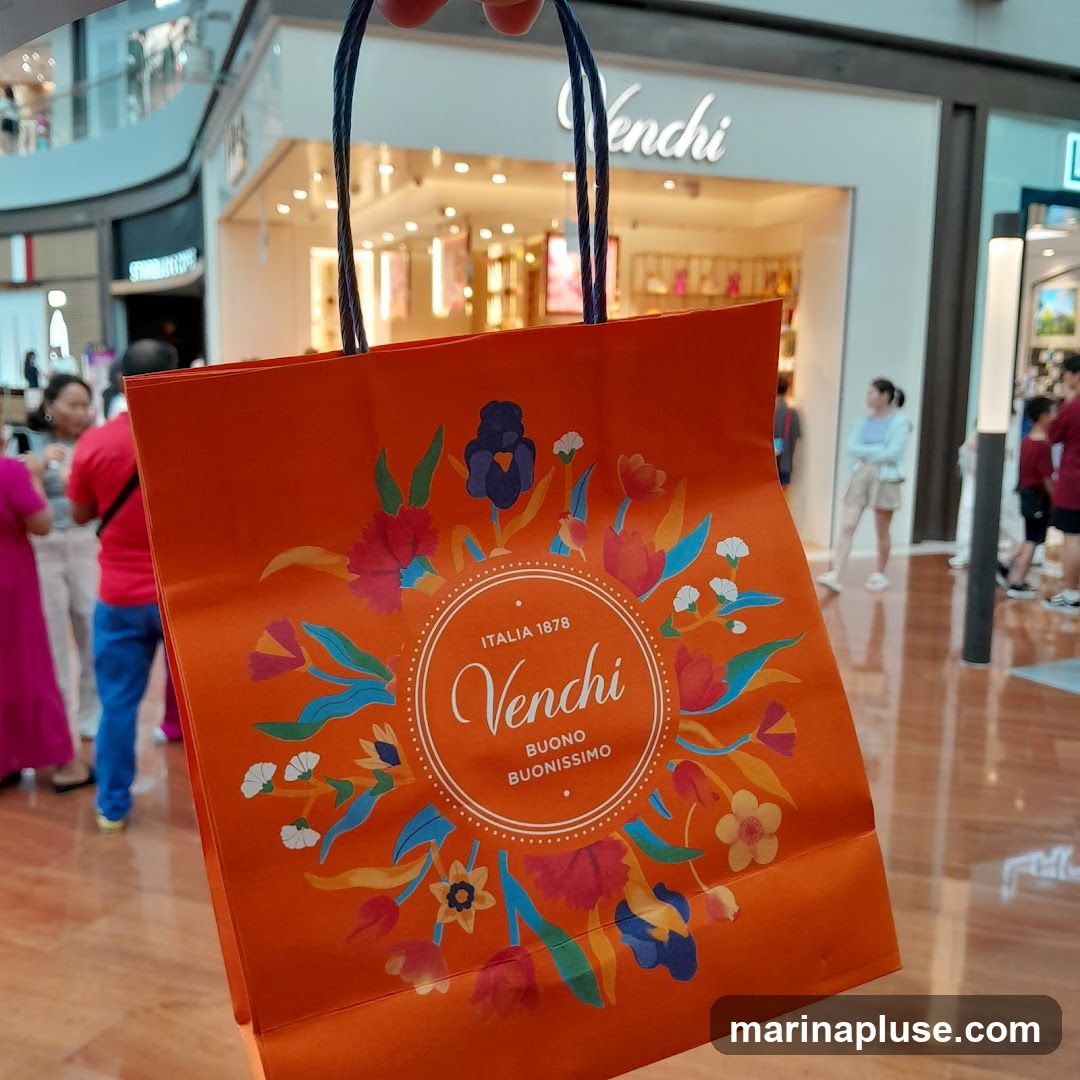 An orange shopping bag with a floral design and the Venchi logo is held in the foreground. In the background, the storefront of a Venchi store is visible, with people browsing inside.