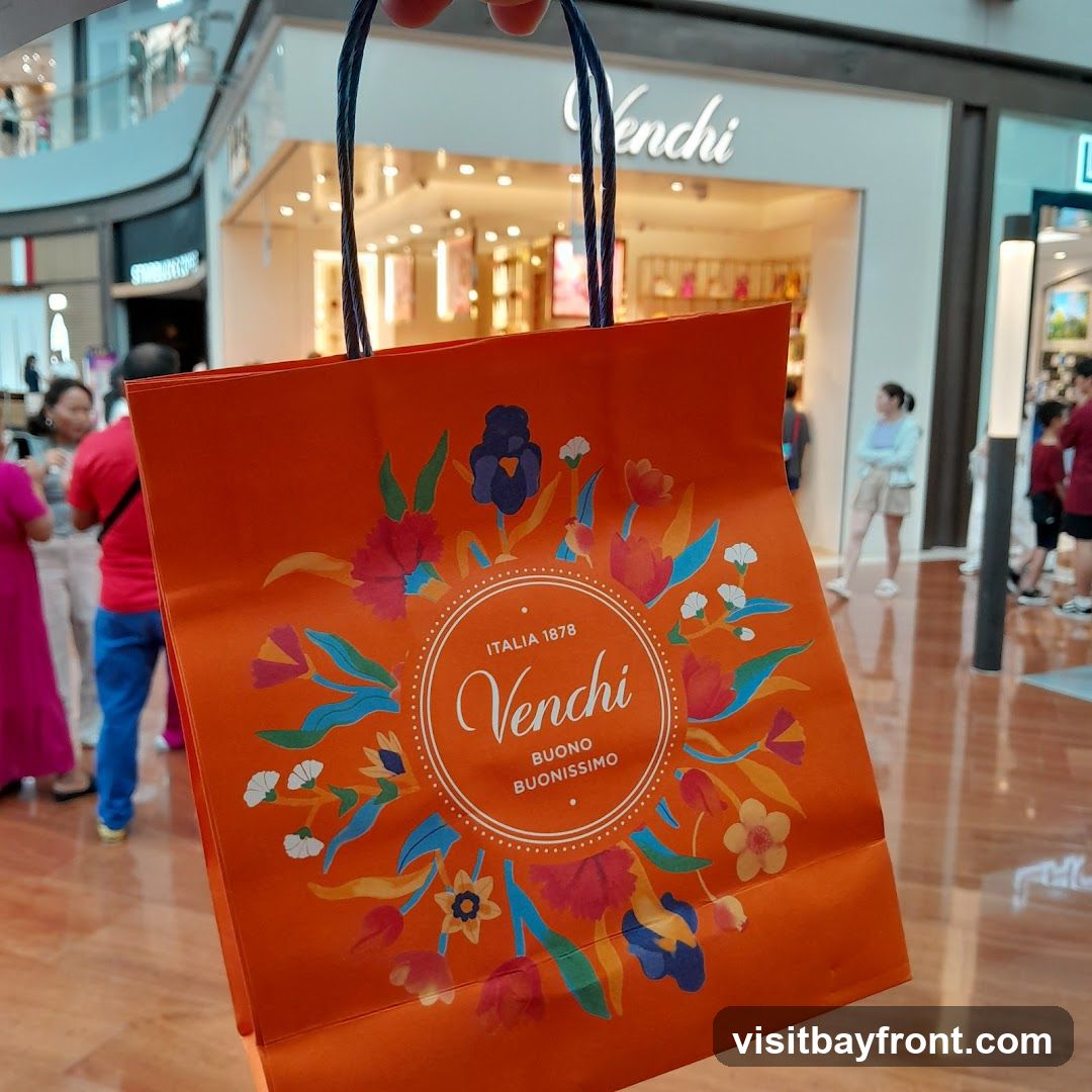 An orange shopping bag with a floral design and the Venchi logo is held in the foreground. In the background, the storefront of a Venchi store is visible, with people browsing inside.