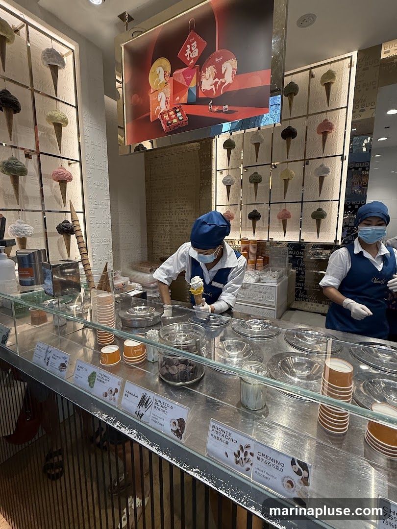 Two employees in blue uniforms and masks are serving ice cream behind a stainless steel counter. The counter displays various flavors of ice cream in metal containers, along with cups and toppings. A wall behind the counter is decorated with artificial ice cream cones in various colors. A large sign above the counter features a festive design with horses and Chinese characters. Several small signs on the counter list ice cream flavors.