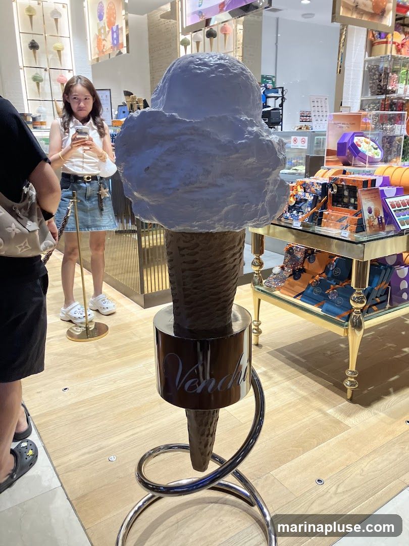 A large, white ice cream cone sculpture is displayed prominently in what appears to be an ice cream shop or confectionery. The cone is dark brown and sits on a metallic stand with the word "Venchi" visible. In the background, a woman is looking at her phone, and shelves display various colorful products, possibly chocolates or other sweets. The flooring is light wood.