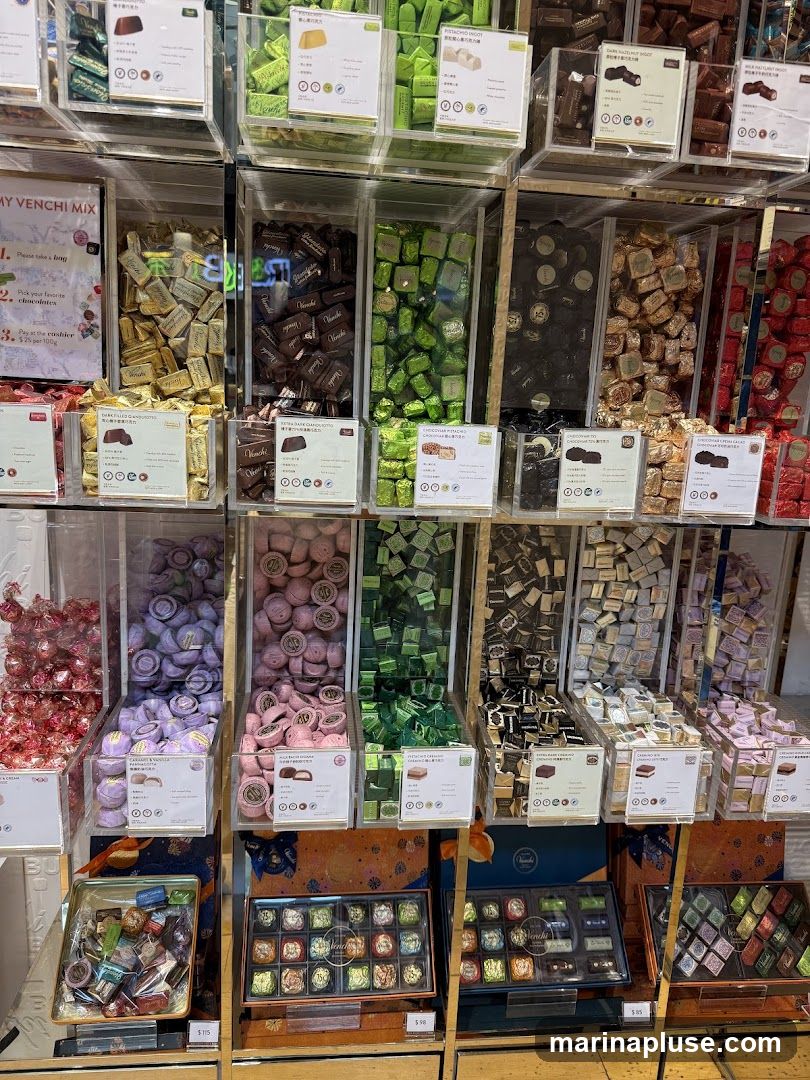 A display of various chocolates in clear plastic bins and boxes. The chocolates are arranged by color and type, including green, purple, pink, brown, and gold wrappers. Some bins have labels with descriptions and prices. Below the bins are boxed assortments of chocolates.