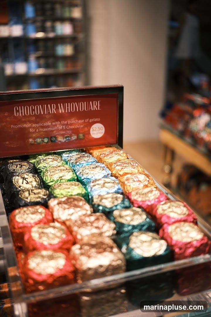 A display of individually wrapped chocolates in various colors, including red, gold, green, blue, and orange. A sign above the chocolates reads "CHOCOVIAR WHOYOUARE" and advertises a promotion applicable with the purchase of gelato, with Chocoviar priced at $2.50 per piece. The background shows shelves stocked with items and a blurred figure of a person.