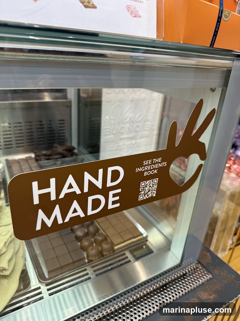 A close-up view of a display case filled with chocolates. A brown sign with white text reads "HAND MADE" and "SEE THE INGREDIENTS BOOK" with a QR code. Inside the case, rows of chocolate bars and chocolate-covered confections are visible.