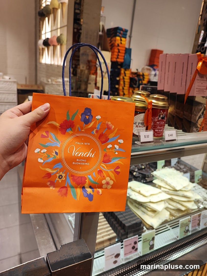 A hand holds an orange Venchi shopping bag with a floral design and the Venchi logo. In the background, a display case shows various chocolates and other confectionery items.
