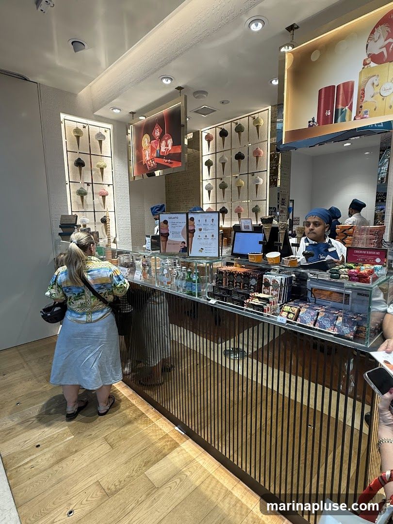 A view of the interior of a gelato shop. A woman stands at the counter, looking at the display. The counter is filled with various products, including chocolates and bottled drinks. Behind the counter, employees are visible. The walls are decorated with images of gelato cones and artwork.