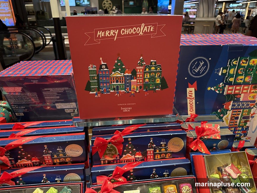A display of Christmas-themed chocolate boxes from Venchi and Bureau Alice. The boxes are decorated with festive illustrations of buildings and Christmas trees. Some boxes are tied with red ribbons. In the background, an escalator and shoppers are visible.