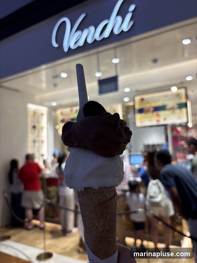 A hand holds a double-scoop gelato cone in front of a Venchi store. The store's illuminated sign is visible above the entrance. Inside, customers are seen ordering and staff are behind the counter.