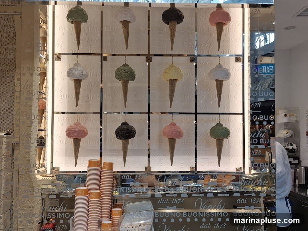 A display of artificial ice cream cones in various flavors, arranged in a grid on a wall. Below the cones is a counter with stacks of paper cups and a mirrored surface reflecting the display and the store interior. The Venchi logo and the phrase "dal 1878" are visible on the counter and the wall. A person in a white uniform is partially visible on the right side of the image.