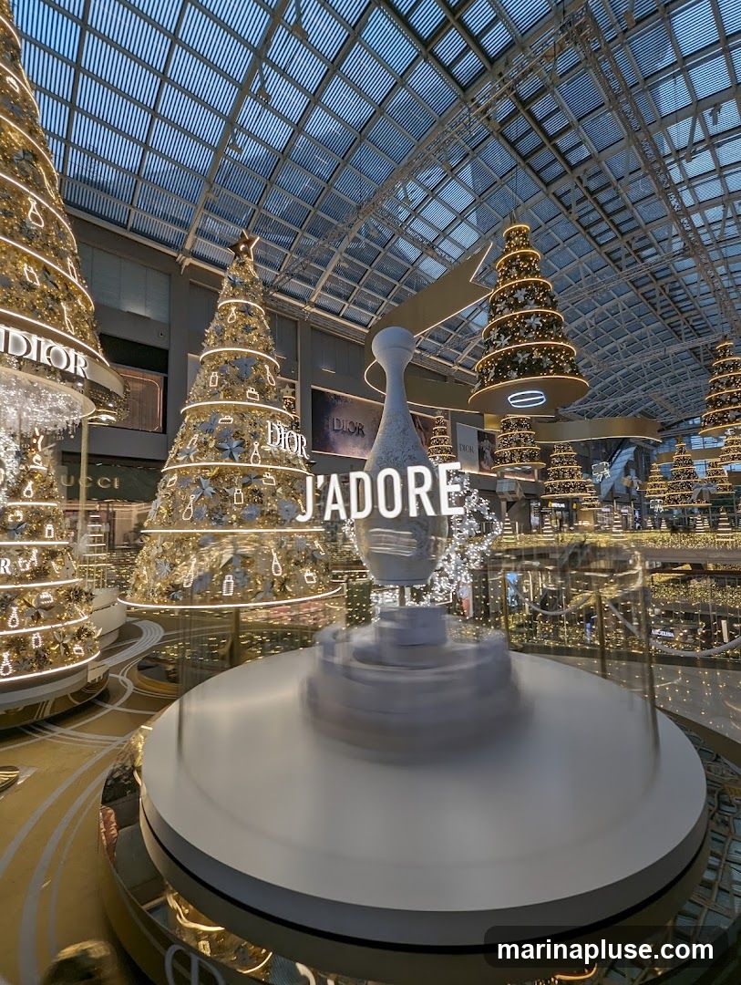 A festive indoor display featuring multiple decorated Christmas trees with lights and ornaments. A large white sculpture with the text "J'ADORE" is prominently displayed in the foreground, rotating on a platform. The background shows a shopping mall interior with more lights and store facades.