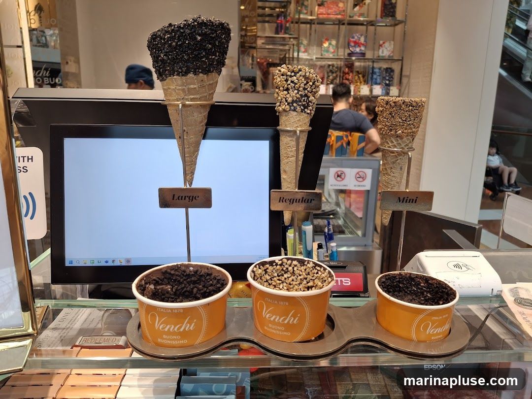 A display of Venchi ice cream cones and cups. There are three cones of different sizes: Large, Regular, and Mini, each topped with various toppings. Below the cones are three cups of ice cream, also from Venchi. The display is set up in front of a computer monitor and behind a glass counter. In the background, shelves are stocked with Venchi products, and people are visible in the distance.