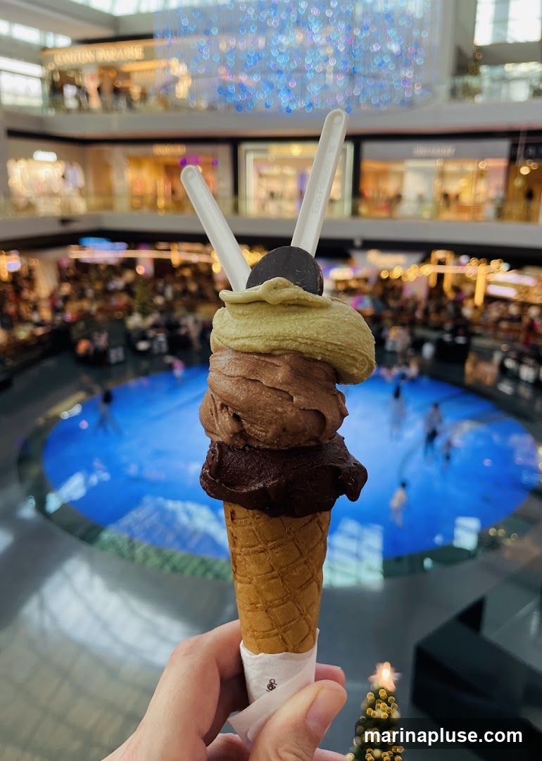 A hand holds a waffle cone with three scoops of ice cream: pistachio, chocolate, and dark chocolate, topped with a chocolate coin and two white spoons. In the background, a brightly lit mall interior is visible with shops, escalators, and a large circular water feature with people walking around it. A small, out-of-focus Christmas tree is in the lower right corner.