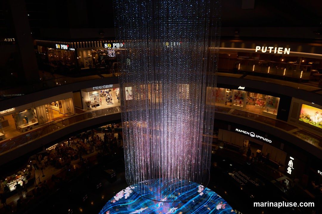 An overhead view of a multi-level indoor shopping mall featuring a large, illuminated art installation in the center. The installation consists of numerous hanging strands of lights that create a shimmering, curtain-like effect, with a circular base displaying a colorful, animated scene. Surrounding the central installation are various shops and restaurants on different levels, including "SO PHO", "FILA KIDS", "PUTIEN", and "RAMEN IPPUDO". People are visible browsing in shops and dining at restaurants.