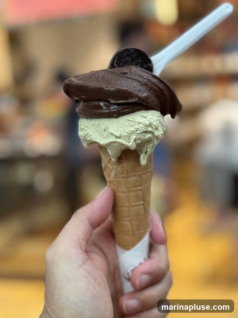 A hand holds a waffle cone with two scoops of gelato. The bottom scoop is a pale green, possibly pistachio, and the top scoop is dark brown, likely chocolate. A piece of an Oreo cookie is on top of the chocolate gelato, and a white plastic spoon is inserted into the cone. The background is blurred, suggesting an indoor setting like an ice cream shop.