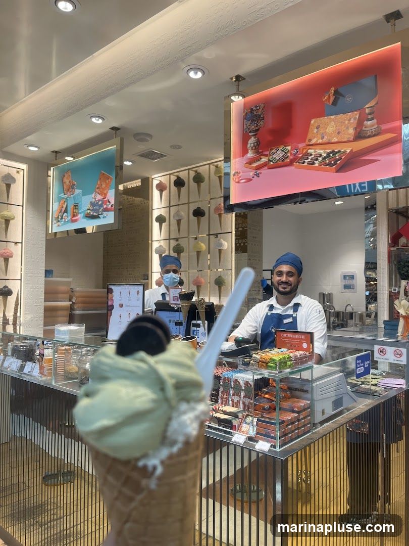 A close-up of a pistachio ice cream cone in the foreground, with two employees behind a counter in the background. The shop interior features displays of chocolates and ice cream. One employee is wearing a mask and the other is smiling at the camera. There are illuminated signs showing boxes of chocolates and ice cream cones.