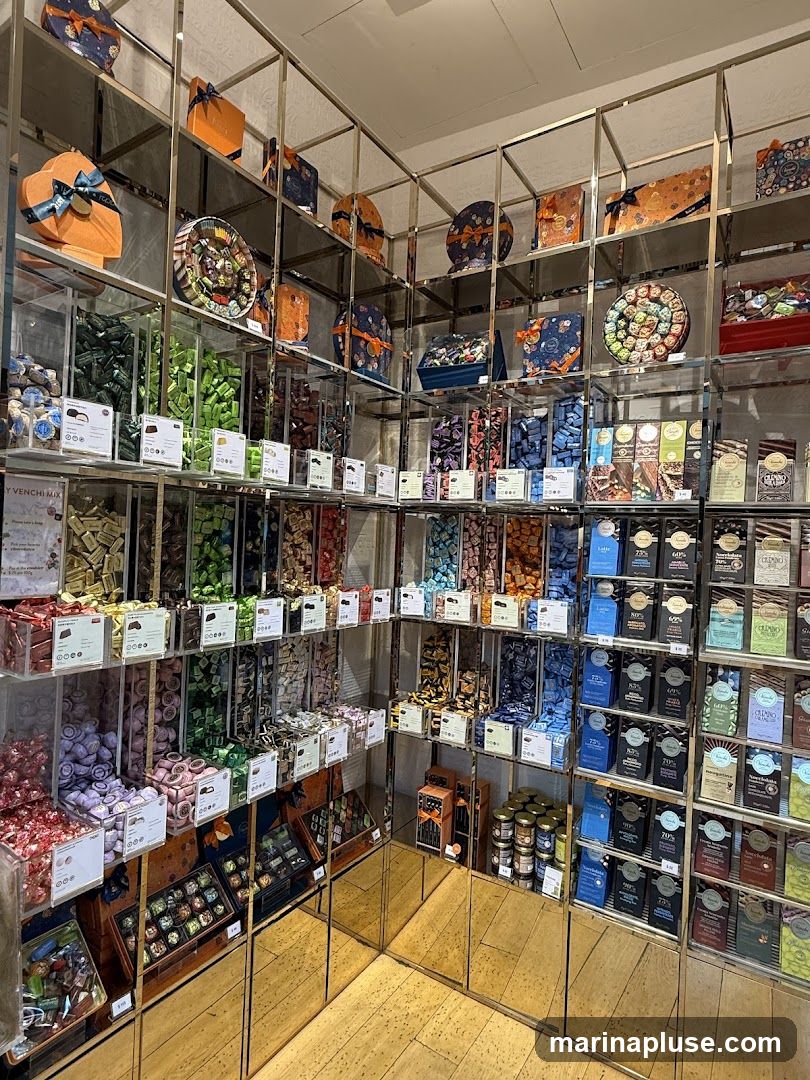 A well-lit interior view of a shop showcasing a wide variety of chocolates and confections. The display features numerous shelves filled with colorful chocolates in boxes, jars, and bulk bins. Some bins contain individually wrapped chocolates in various colors, while others hold assortments. Packaged chocolate bars are neatly arranged on shelves, with some displaying percentages like 70% and 80%. Decorative boxes with ribbons are also visible on higher shelves. The lower shelves and display cases have mirrored surfaces, reflecting the products and the wooden floor. Small labels are placed in front of many bins, likely indicating flavors or prices.