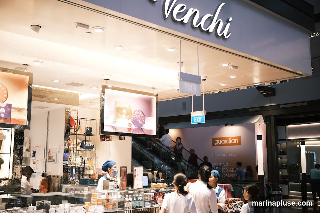 A view of the Venchi store, with its prominent signage visible above the entrance. Inside, staff members are serving customers behind a display counter filled with chocolates and other treats. Various products are displayed on shelves and in illuminated cases. An escalator is visible in the background, along with an "EXIT" sign and a large advertisement for "guardian".