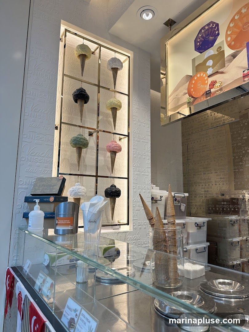 A display of artificial ice cream cones in various flavors, presented on shelves. Below, a glass counter shows ice cream cones, containers, and some small printed menus. The background features the Venchi logo and branding.